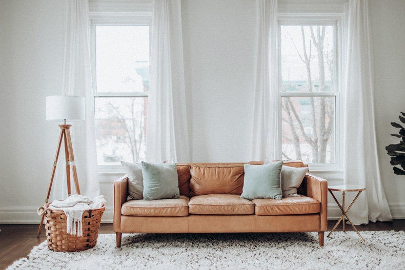 a white minimalist living room containing a brown leather couch with white curtains behind, a rattan basket, a wooden floor lamp